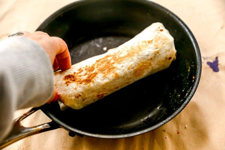 A hand holds a toasted burrito in a black skillet on a light-colored surface.