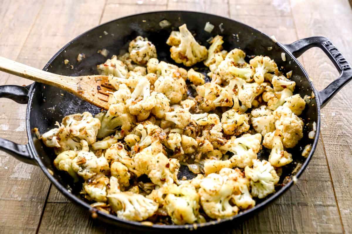 Cauliflower florets sautéing in a black skillet with onions and seasonings, stirred with a wooden spoon on a wooden surface.
