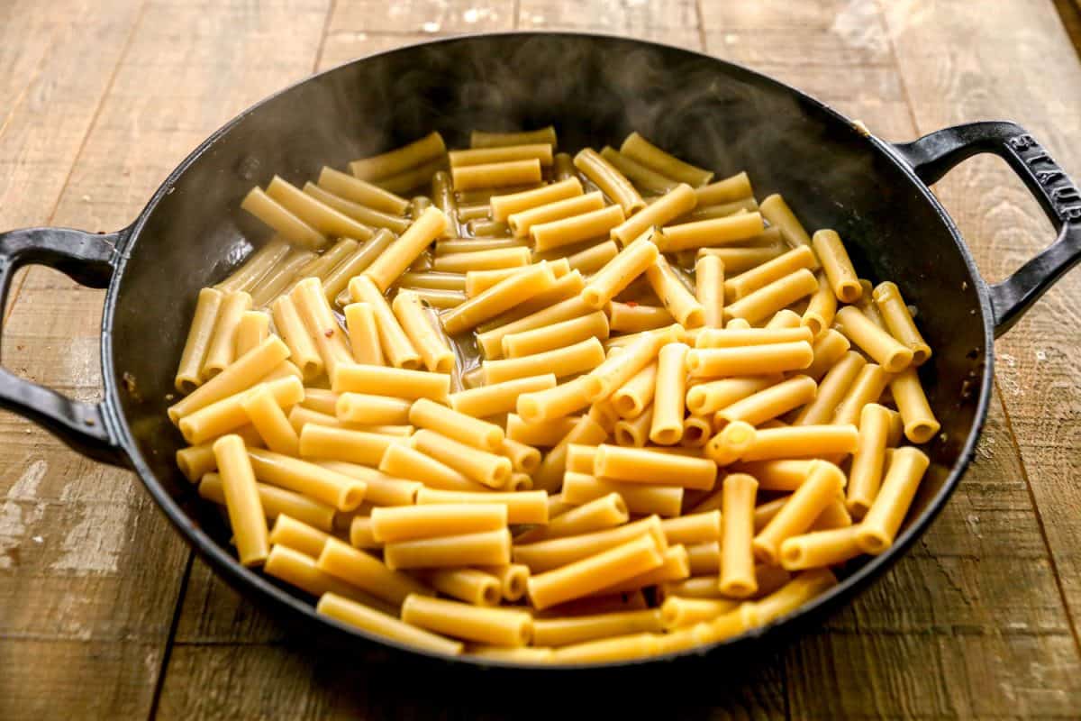 Cooked ziti pasta in a black cast iron pan on a wooden surface, with some steam rising.