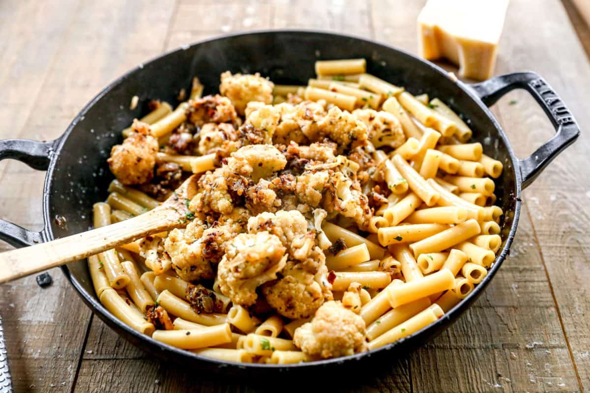 Pasta with cauliflower in a black skillet on a wooden table, stirred with a wooden spoon.