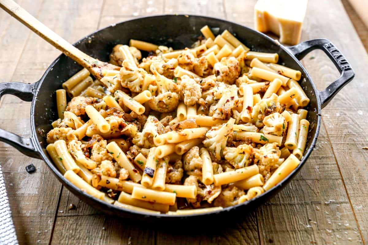 Pasta with cauliflower and herbs in a black skillet on a wooden table, stirred with a wooden spoon.