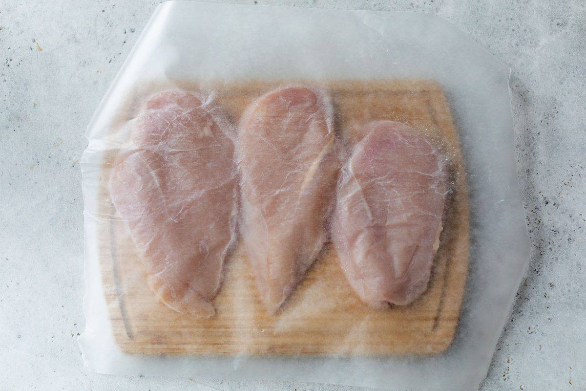 Three raw chicken breasts on a wooden cutting board, covered with a sheet of wax or parchment paper.