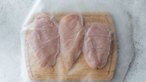 Three raw chicken breasts on a wooden cutting board, covered with a sheet of wax or parchment paper.