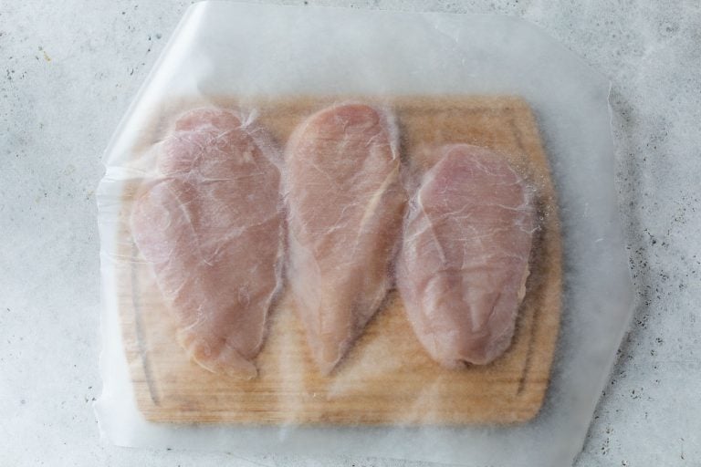 Three raw chicken breasts on a wooden cutting board, covered with a sheet of wax or parchment paper.