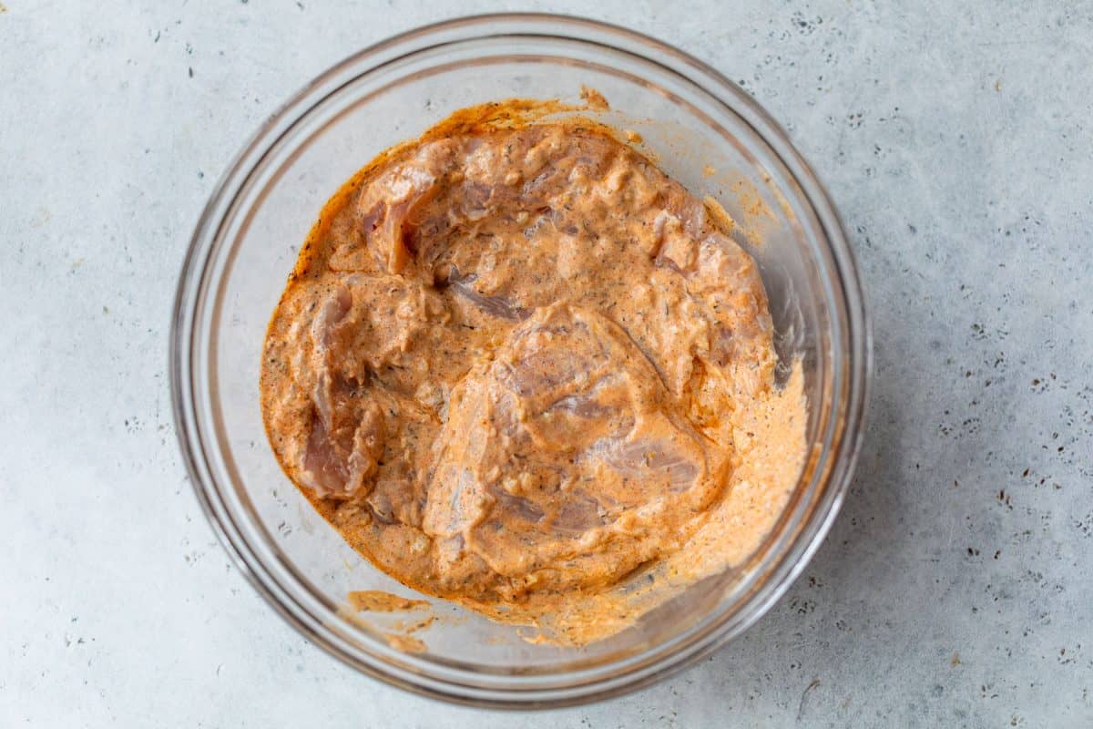 Raw chicken marinating in a spiced yogurt mixture in a clear glass bowl on a light gray surface.