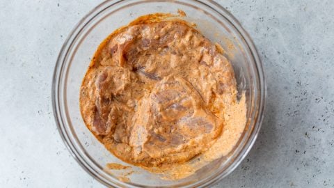 Raw chicken marinating in a spiced yogurt mixture in a clear glass bowl on a light gray surface.