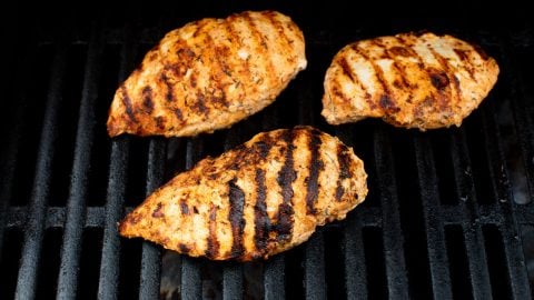 Three seasoned chicken breasts with grill marks cooking on a black grill grate.