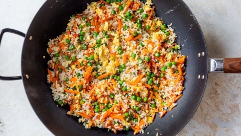 A wok filled with fried rice, peas, carrots, and scrambled eggs on a light-colored surface.
