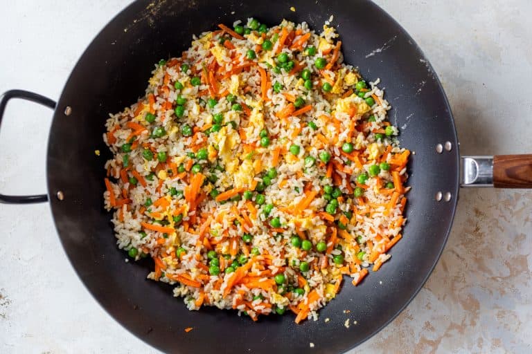 A wok filled with fried rice, peas, carrots, and scrambled eggs on a light-colored surface.