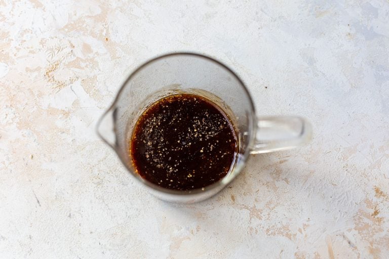 Clear glass measuring cup with dark fried rice sauce and visible pepper specks on a light textured surface.