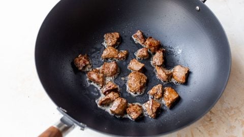 Chunks of browned beef sizzling in oil inside a black wok on a light-colored surface.