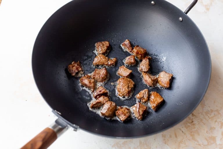 Chunks of browned beef sizzling in oil inside a black wok on a light-colored surface.