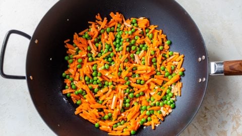 Sliced carrots and green peas being stir-fried in a black wok on a light-colored surface.