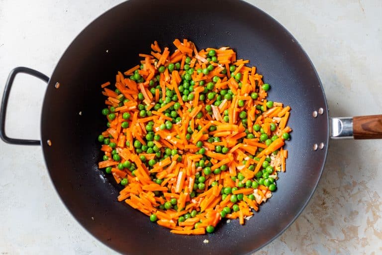 Sliced carrots and green peas being stir-fried in a black wok on a light-colored surface.