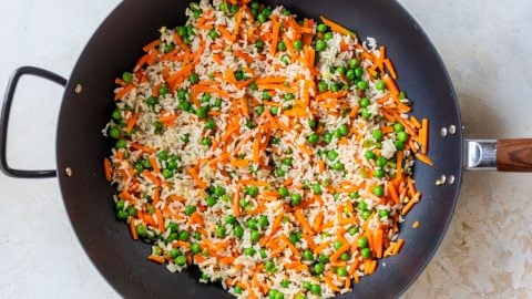A black skillet filled with fried rice, peas, and carrot strips on a light-colored surface.