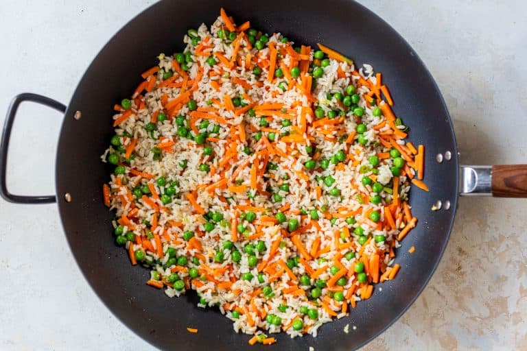 A black skillet filled with fried rice, peas, and carrot strips on a light-colored surface.