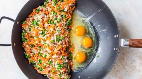 A wok with fried rice, carrots, peas on one side and three raw eggs on the other side, ready to be mixed.