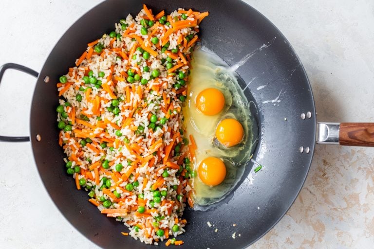 A wok with fried rice, carrots, peas on one side and three raw eggs on the other side, ready to be mixed.