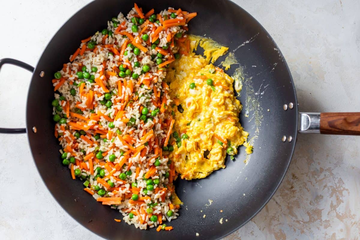 Wok with fried rice, peas, carrots on one side and scrambled eggs on the other, ready to be mixed.