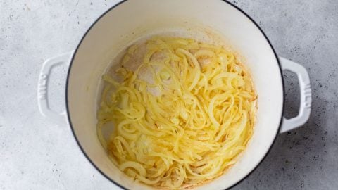 Sliced onions cooking and softening in a white Dutch oven on a light gray surface.