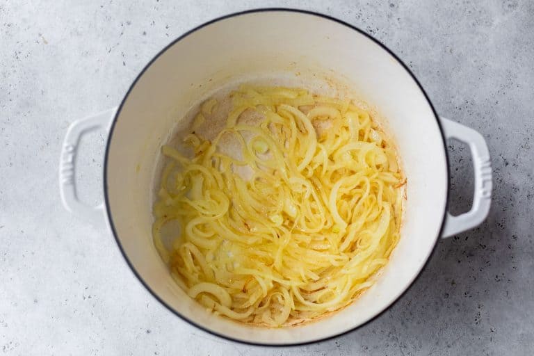 Sliced onions cooking and softening in a white Dutch oven on a light gray surface.