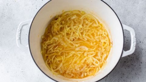 Sliced onions caramelizing in a white Dutch oven on a light gray surface.
