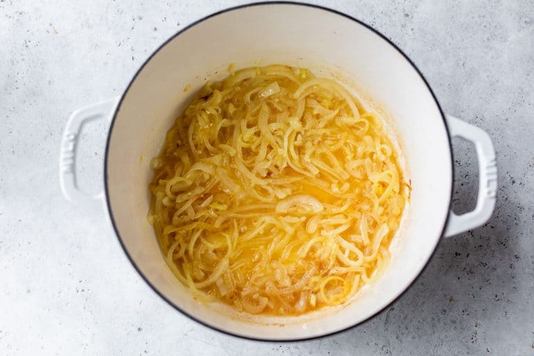 Sliced onions caramelizing in a white Dutch oven on a light gray surface.