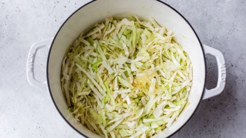 White pot filled with thinly sliced cabbage on a light gray surface, ready for cooking.