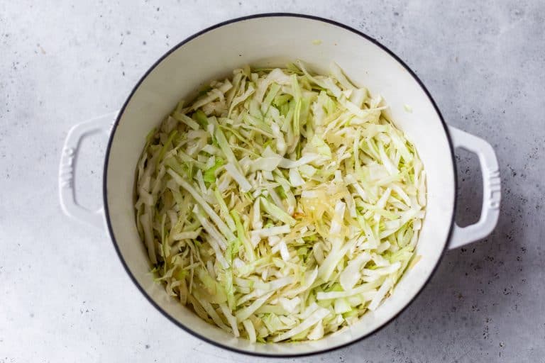 White pot filled with thinly sliced cabbage on a light gray surface, ready for cooking.