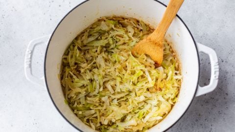 Shredded steamed cabbage in a white pot with a wooden spoon on a light gray surface.