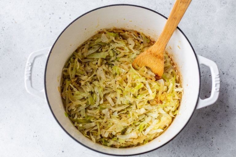 Shredded steamed cabbage in a white pot with a wooden spoon on a light gray surface.