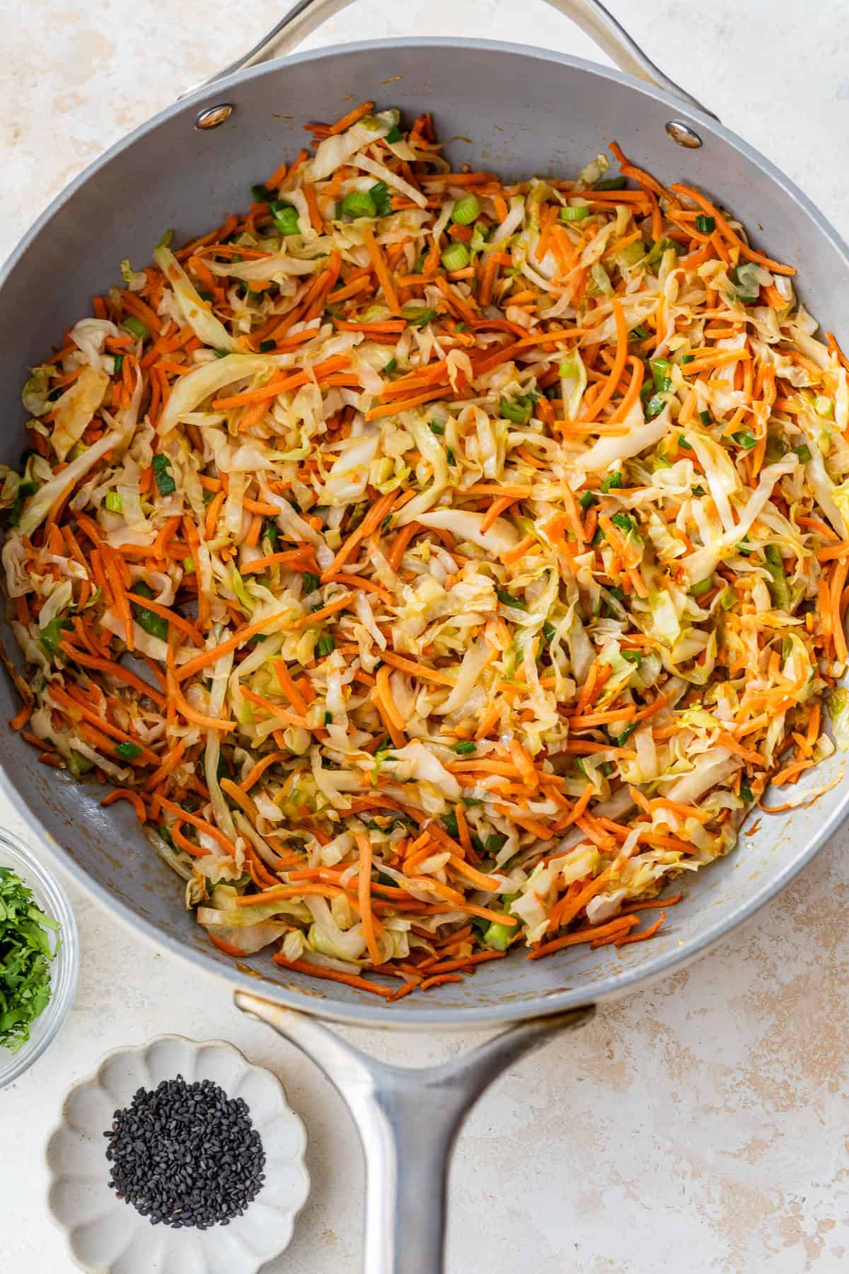 Sautéed cabbage and carrots with green onions in a large skillet, next to bowls of cilantro and black sesame seeds.