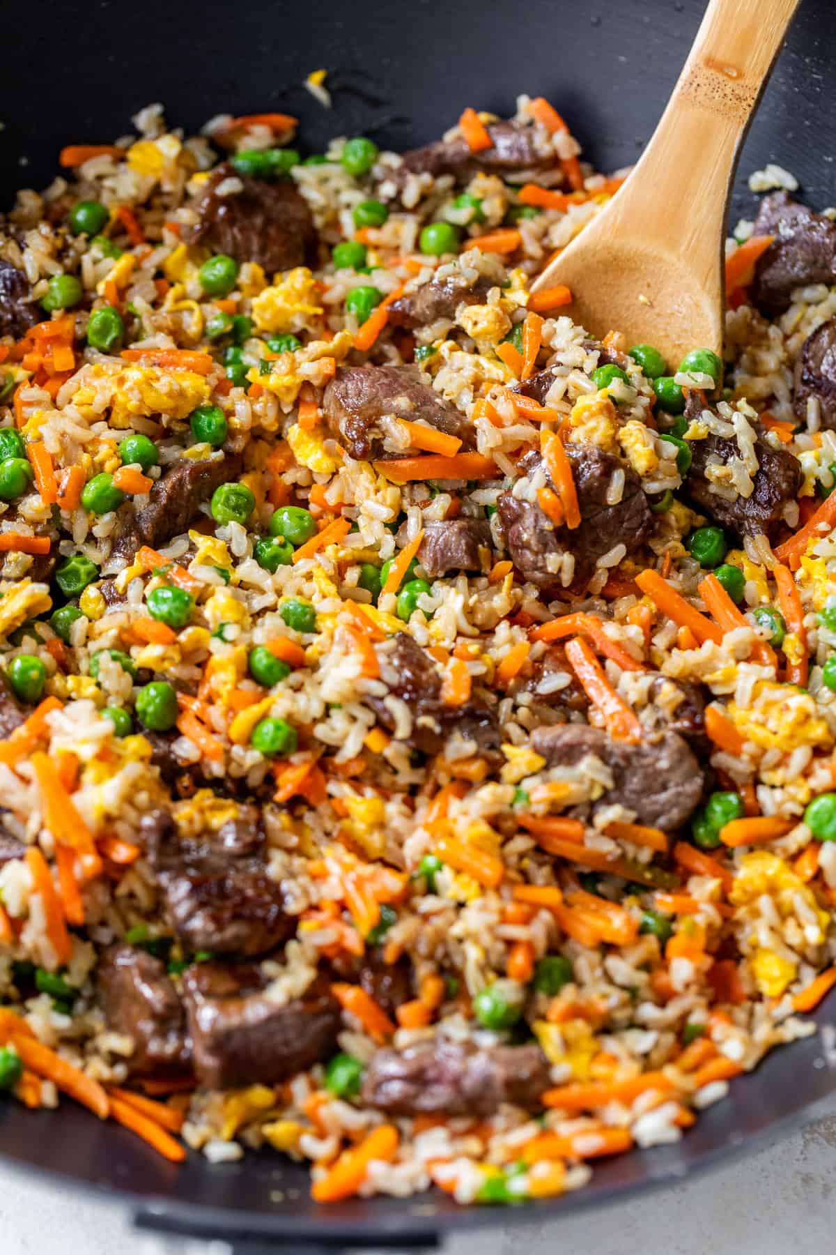 A close-up of steak fried rice with peas, carrots, scrambled eggs, and a wooden spoon in a black pan.