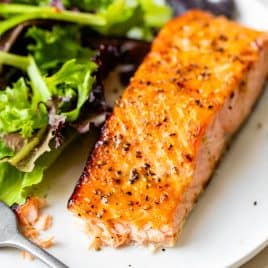 A piece of cooked salmon with black pepper beside a serving of mixed green salad on a white plate.
