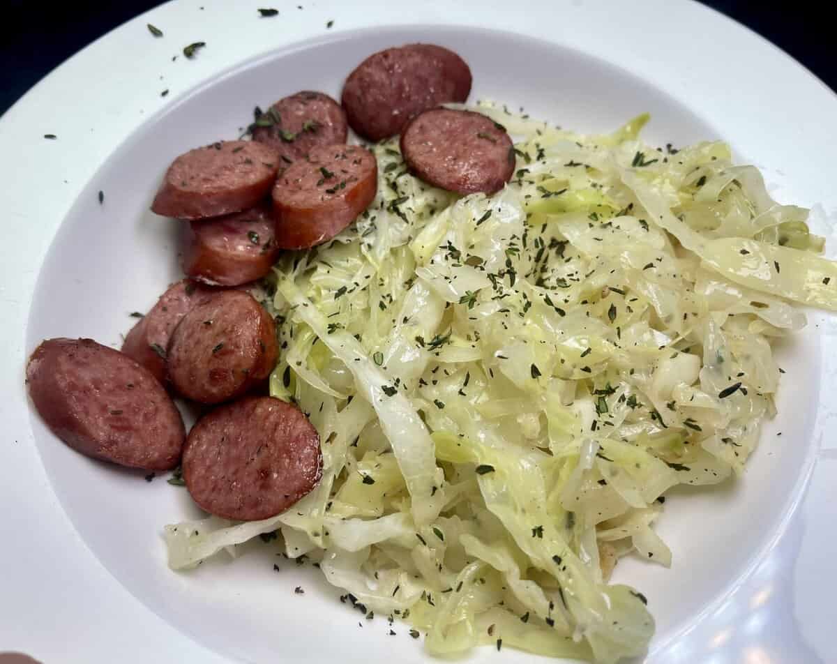 Sliced sausage and cooked cabbage garnished with herbs, served on a white plate.