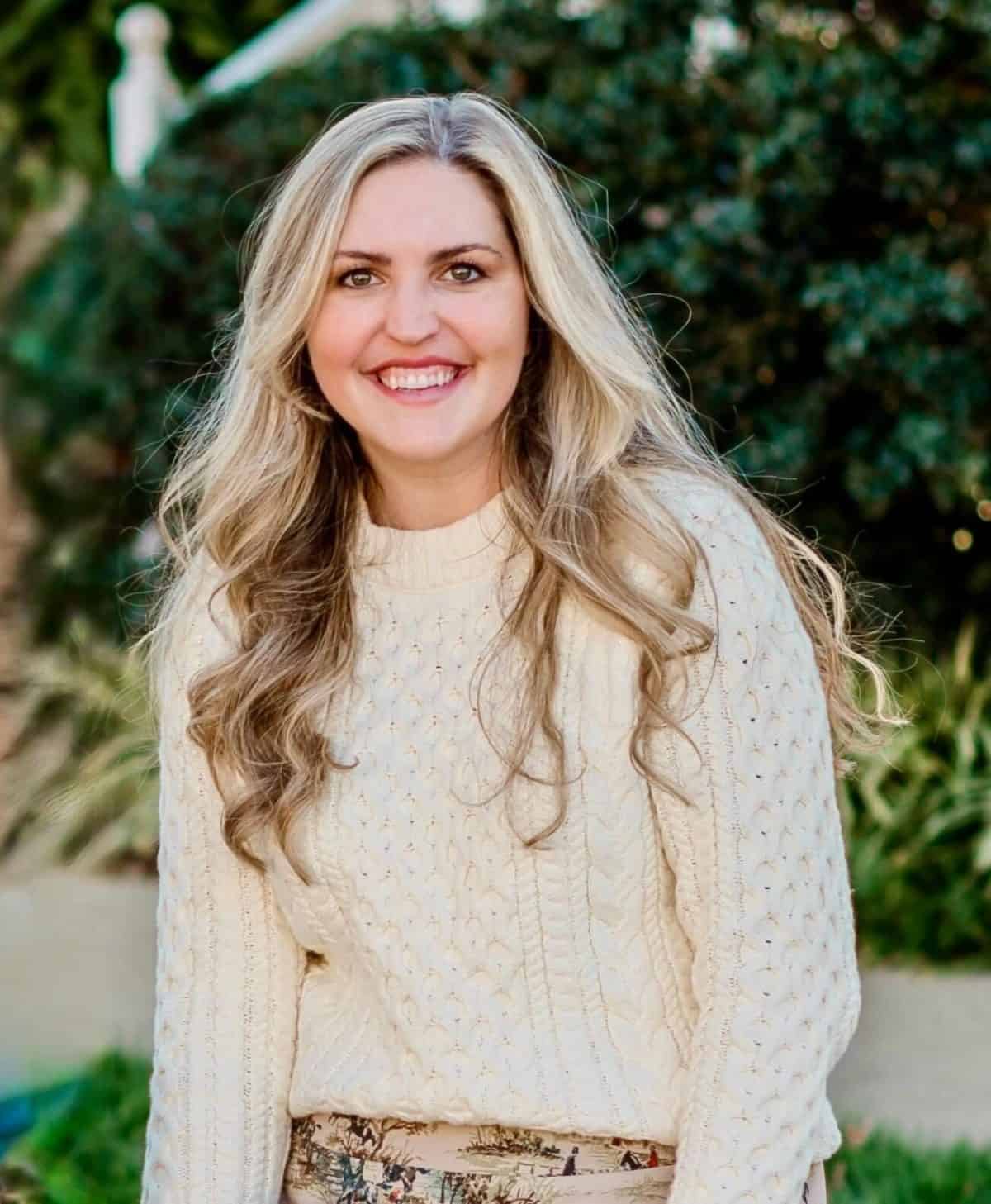 Smiling woman with long blonde hair wearing a cream cable-knit sweater, outdoors with greenery in the background.