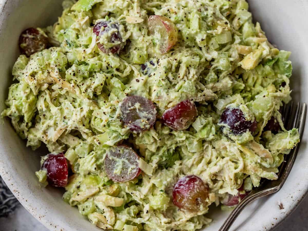 A bowl of avocado chicken salad with grapes, and celery, with a fork and a halved avocado beside the bowl.