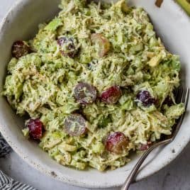 A bowl of avocado chicken salad with grapes, and celery, with a fork and a halved avocado beside the bowl.