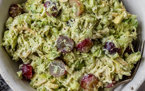 A bowl of avocado chicken salad with grapes, and celery, with a fork and a halved avocado beside the bowl.