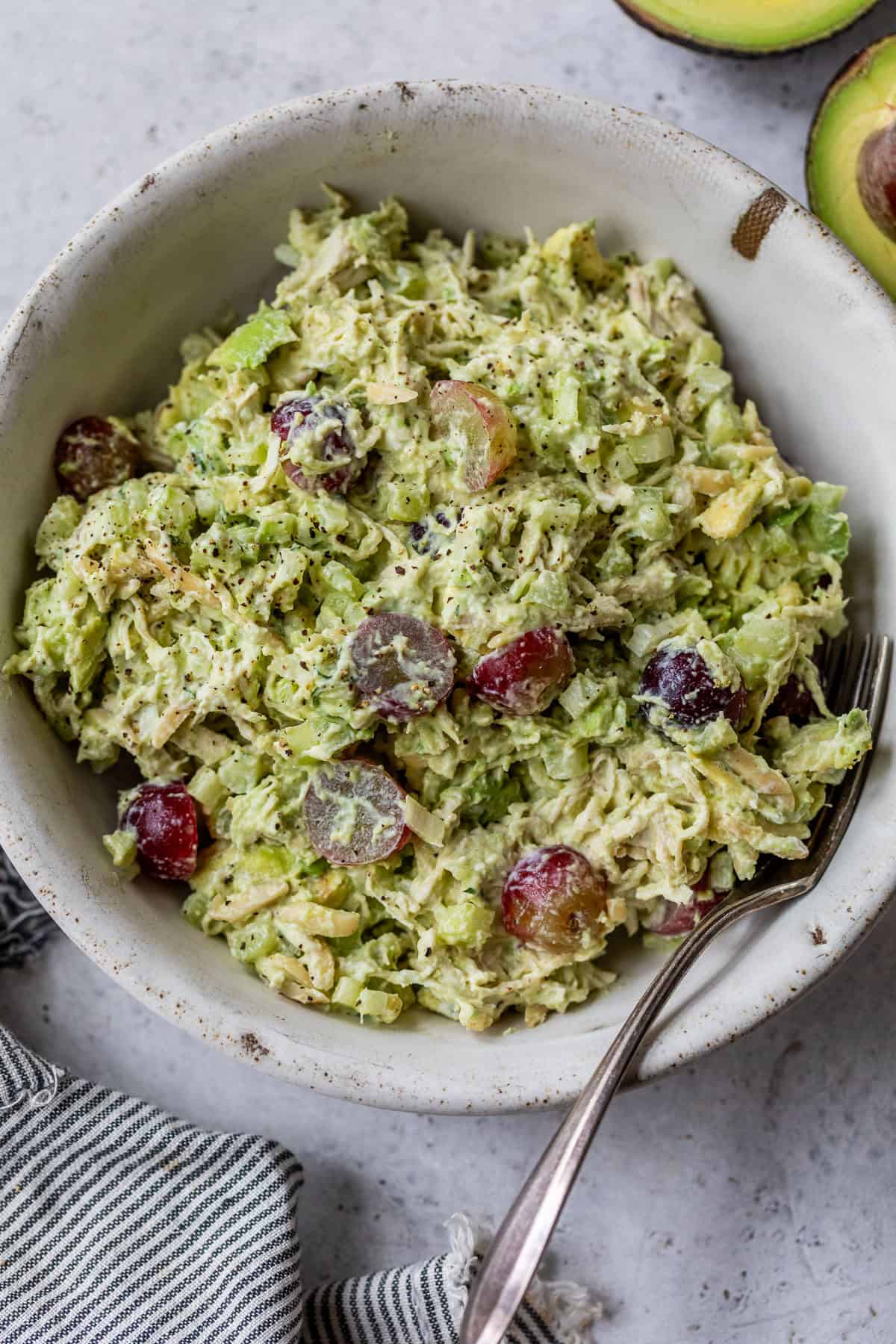 A bowl of avocado chicken salad with grapes, and celery, with a fork and a halved avocado beside the bowl.