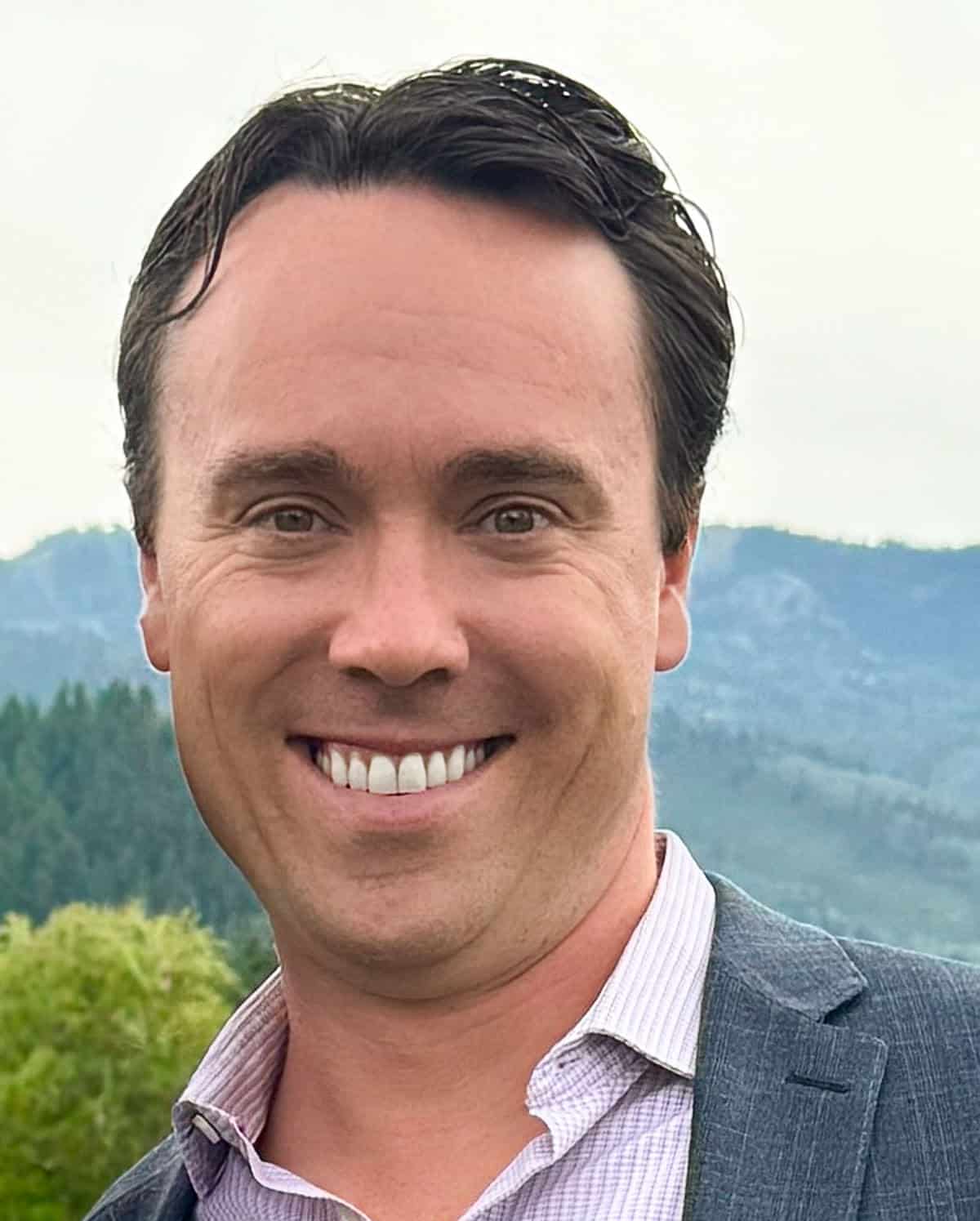 A man in a suit smiling outdoors with mountains and trees in the background.