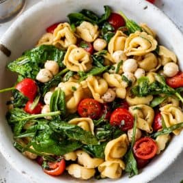 Bowl of tortellini pasta salad with spinach, cherry tomatoes, mozzarella balls, and herbs on a light surface.
