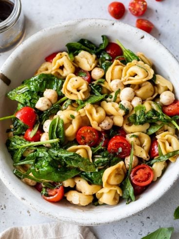 Bowl of tortellini pasta salad with spinach, cherry tomatoes, mozzarella balls, and herbs on a light surface.