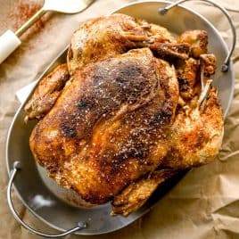 A crockpot whole chicken with crispy, seasoned skin sits in a metal roasting pan on brown parchment paper.