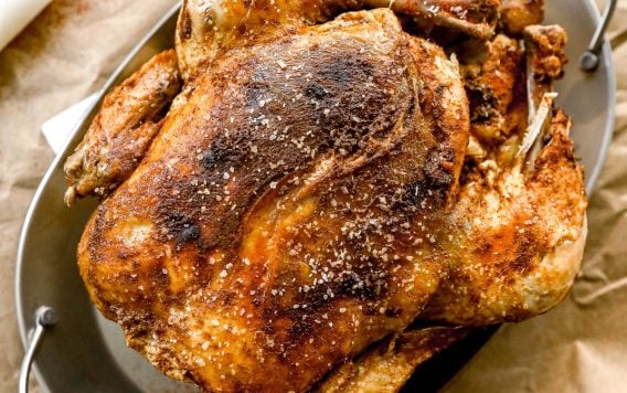 A crockpot whole chicken with crispy, seasoned skin sits in a metal roasting pan on brown parchment paper.