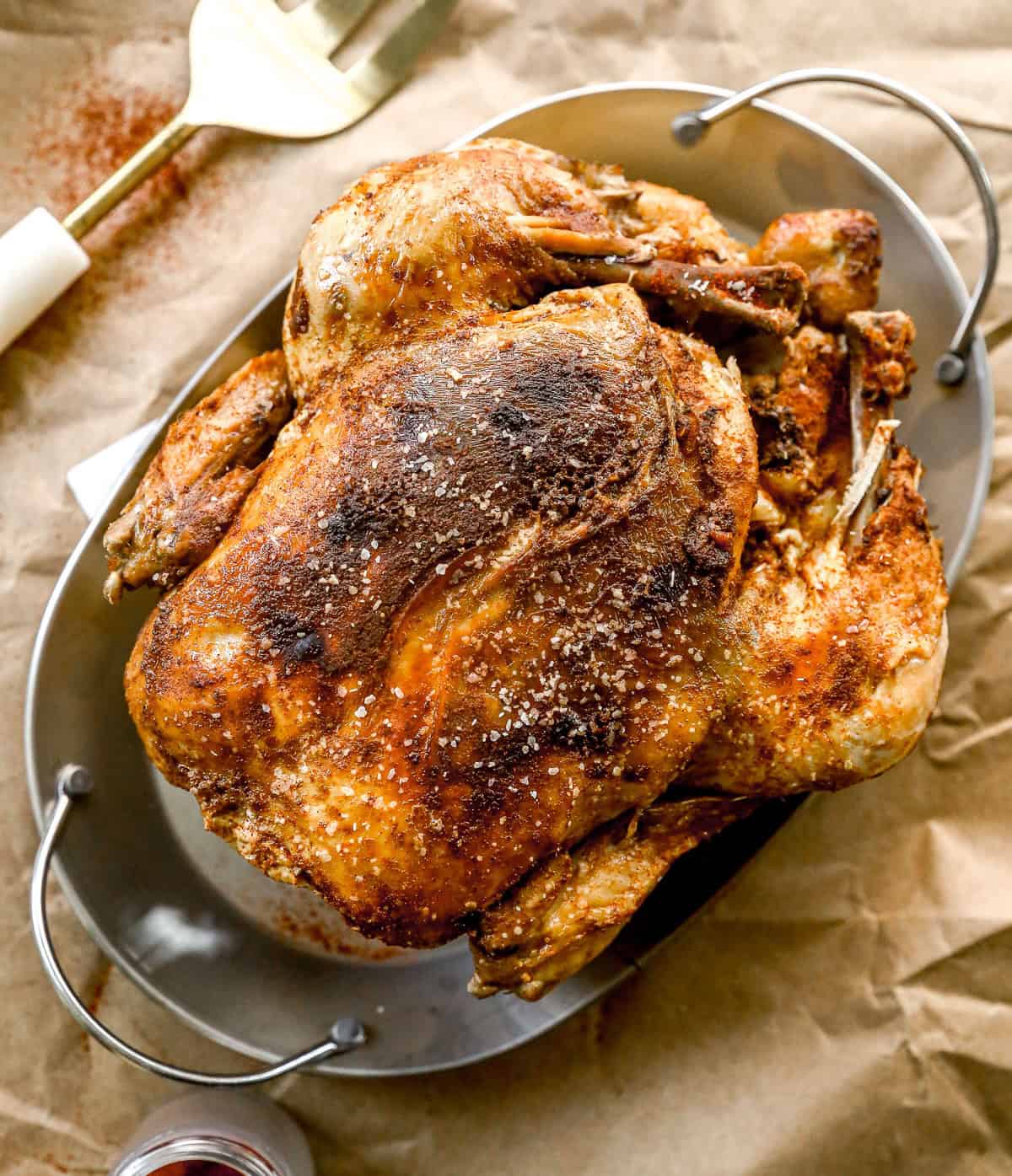 A crockpot whole chicken with crispy, seasoned skin sits in a metal roasting pan on brown parchment paper.