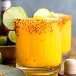 A glass of mango margarita with a chili powder rim, ice, and lime slices, sitting on a wooden table.