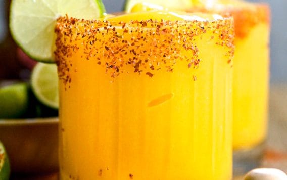 A glass of mango margarita with a chili powder rim, ice, and lime slices, sitting on a wooden table.