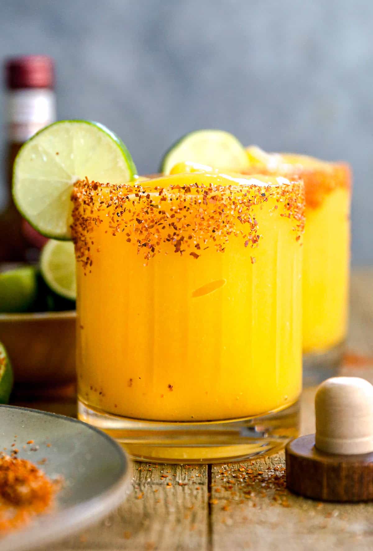 A glass of mango margarita with a chili powder rim, ice, and lime slices, sitting on a wooden table.