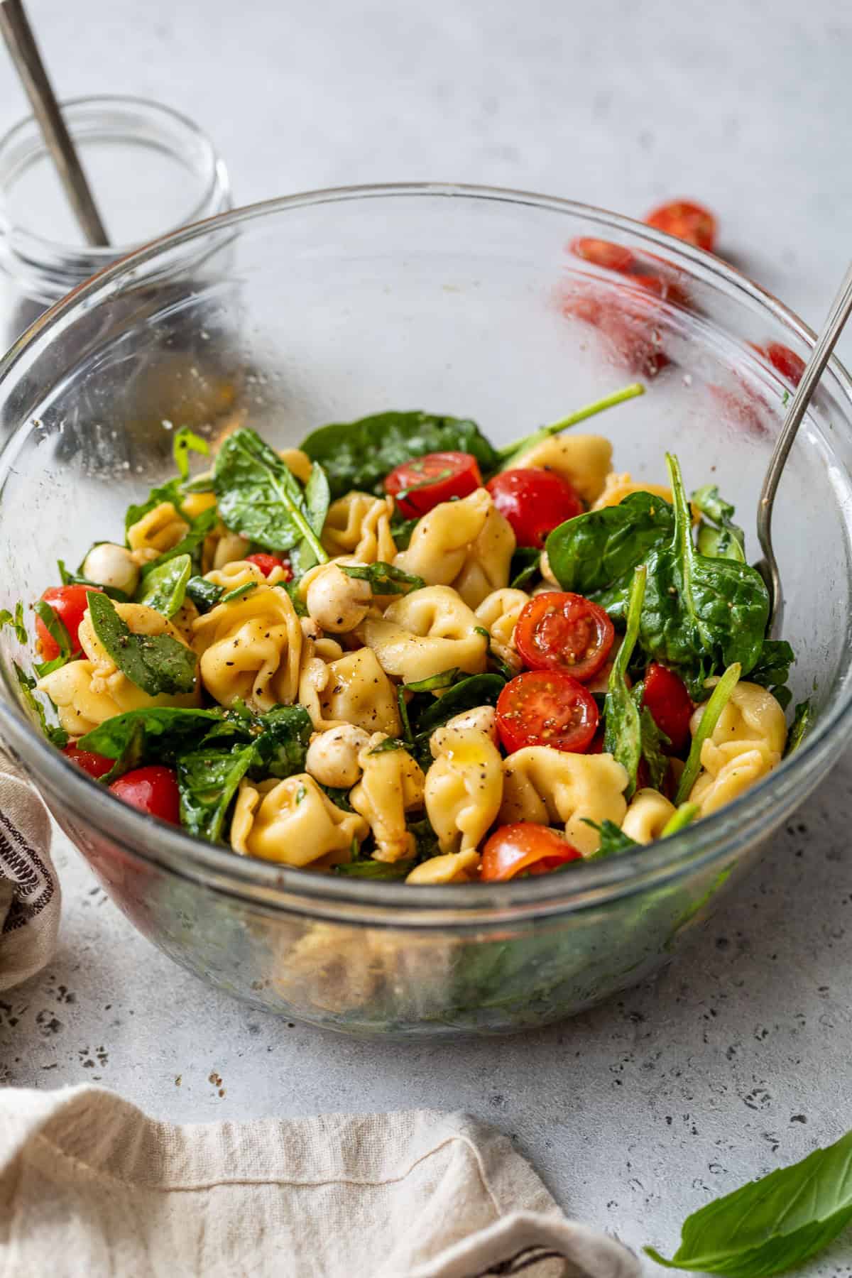Glass bowl of caprese pasta salad with spinach, cherry tomatoes, chickpeas, and herbs, with serving utensils inside.